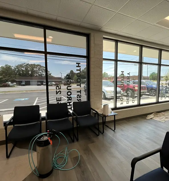 Waiting area inside a commercial office with large tinted windows, dark chairs, and a window tinting installation setup, including a spray tank and hose.