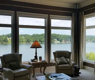Interior of a cozy living room with large picture windows offering a scenic view of a lake and tree-lined shore. The room features two armchairs, a table with a lamp, and soft natural light filling the space.