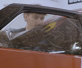 Technician installing window tint film on the driver-side window of a red car, with water and air bubbles visible under the film during application.