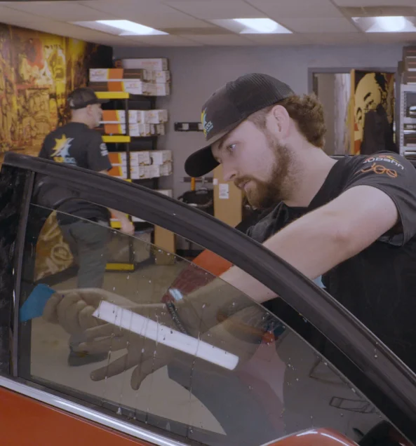 Window tint technician carefully applying film to a car door window using a squeegee inside a workshop, with shelves of tint rolls and tools visible in the background.