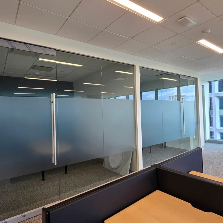 Modern office with floor-to-ceiling glass walls featuring frosted privacy film installed across the center, dividing private rooms from open workspaces.