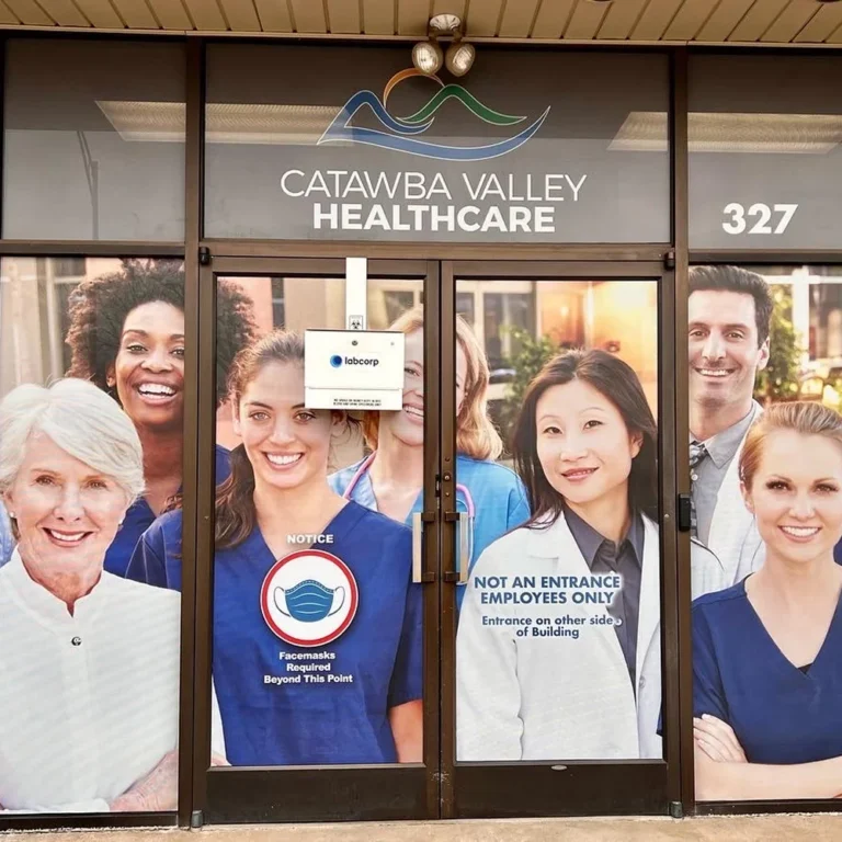 Catawba Valley Healthcare building entrance with full-window vinyl graphics of diverse medical staff, including a facemask requirement notice and directional signage for employees.