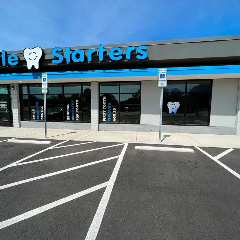 Storefront of Smile Starters dental office with large tinted windows featuring white and blue vinyl lettering and graphics, viewed from a freshly striped parking lot.