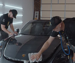 Two technicians applying paint protection film to the hood of a black Porsche inside a detailing shop.