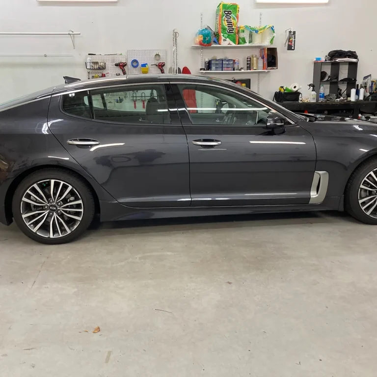 A dark gray Kia Stinger sedan parked inside an auto detailing shop, with polished multi-spoke alloy wheels and a clean, glossy exterior.