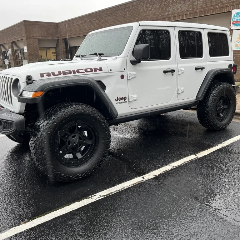 A white Jeep Wrangler Rubicon with large off-road tires and black wheels parked on a wet asphalt lot in front of a brick commercial building on a rainy day.