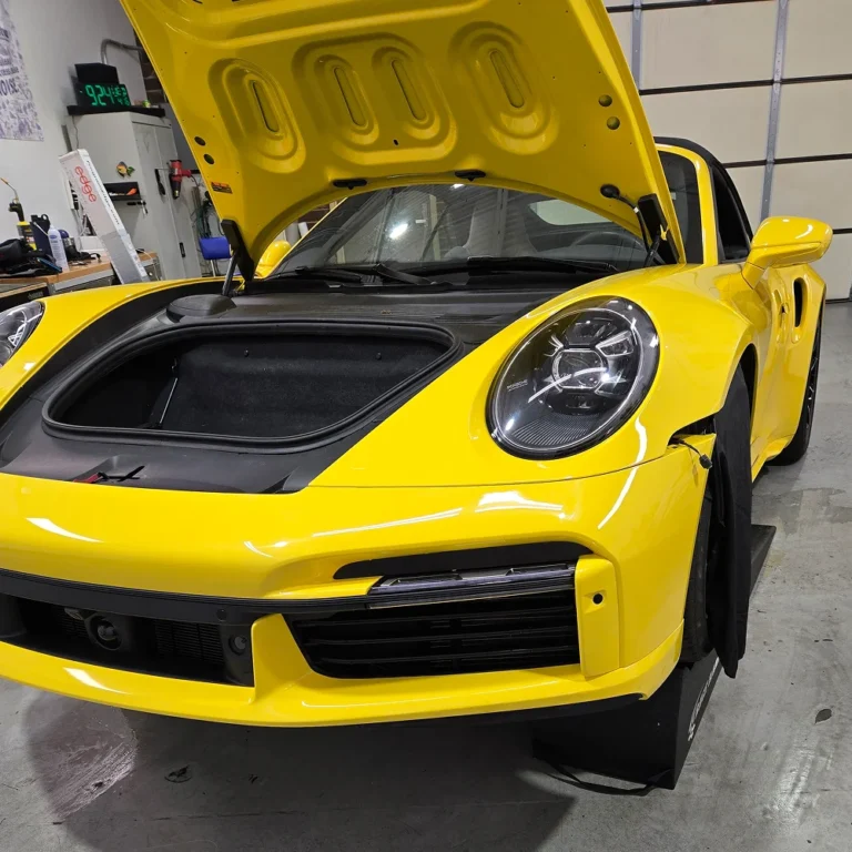 A bright yellow Porsche 911 with its front trunk open inside an automotive shop, showing the empty frunk compartment and detailed LED headlight design.