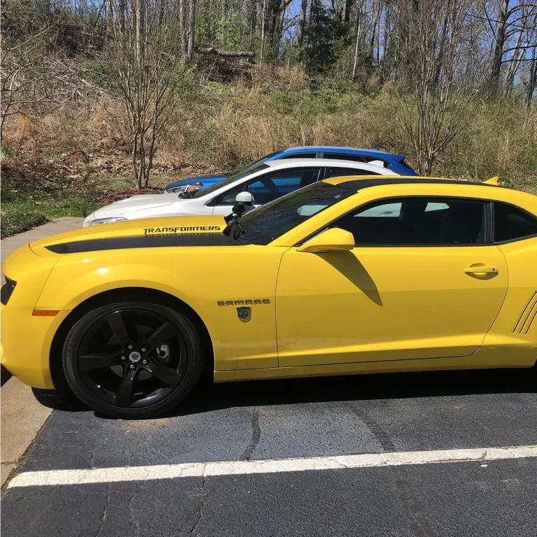 Bright yellow Chevrolet Camaro with tinted windows and black “Transformers” decals, parked in a sunlit lot next to other vehicles.