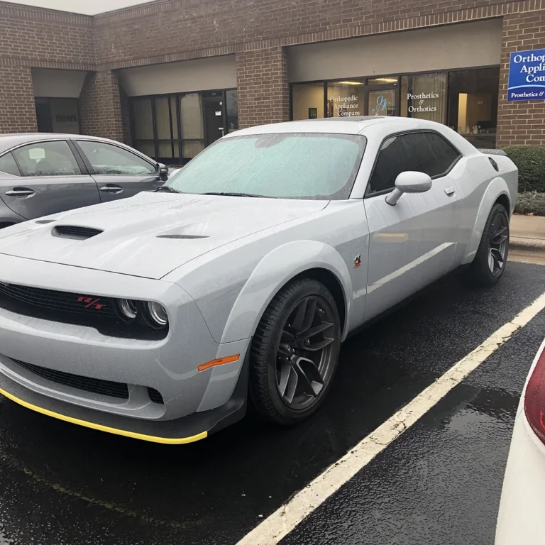 A light gray Dodge Challenger R/T with a widebody kit and yellow splitter guards parked on a wet asphalt lot in front of a brick medical office building.