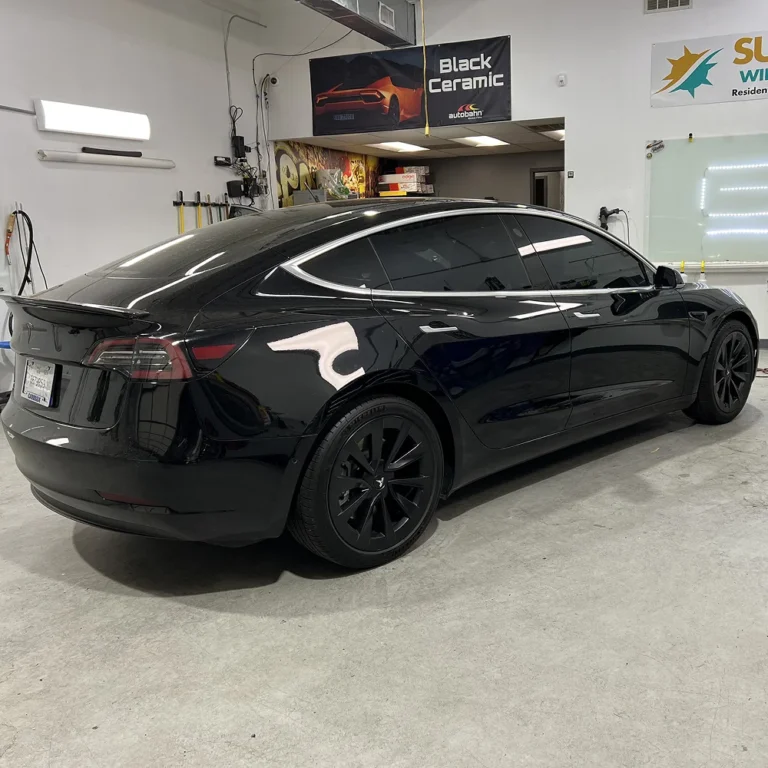 A black Tesla Model 3 with tinted windows and black wheels parked inside a detailing shop, with banners advertising "Black Ceramic" and "Sun Window Tinting" in the background.