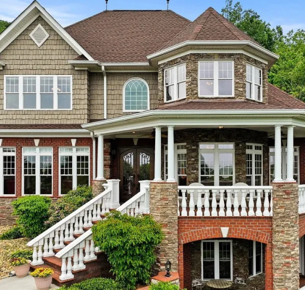 A large two-story house featuring a mix of stone, brick, and shake siding, with a wraparound porch, white balustrade railings, and a turret-style upper corner. A grand staircase leads to the front door, surrounded by manicured shrubs and potted plants.