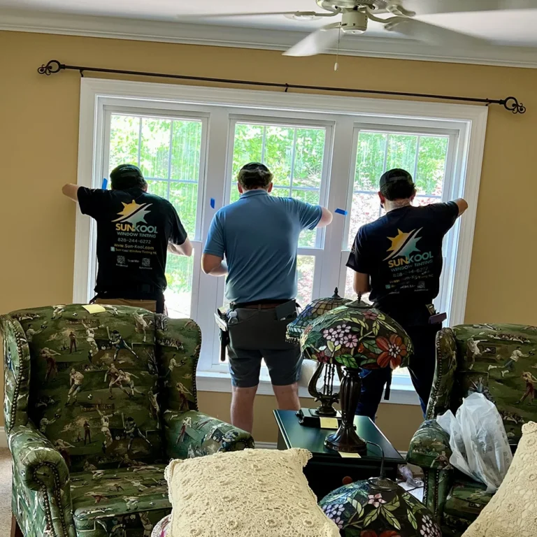 Three men from Sun Kool Window Tinting install film on large living room windows, using tape to secure the material. The room is decorated with floral stained glass lamps and hunting-themed armchairs.