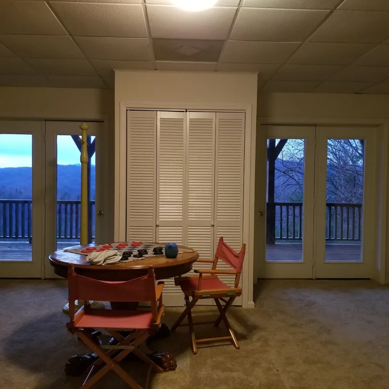 Interior view of a room with a round wooden table and red canvas chairs, showing large glass doors with privacy window tint and a scenic mountain view in the background.