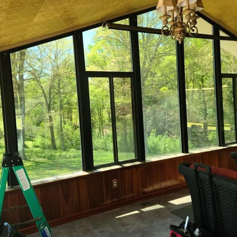 A sunroom with large floor-to-ceiling windows framed in black, offering a clear view of a lush green forest. The interior features wood paneling, a vintage chandelier, and a glimpse of wicker furniture and a green ladder.