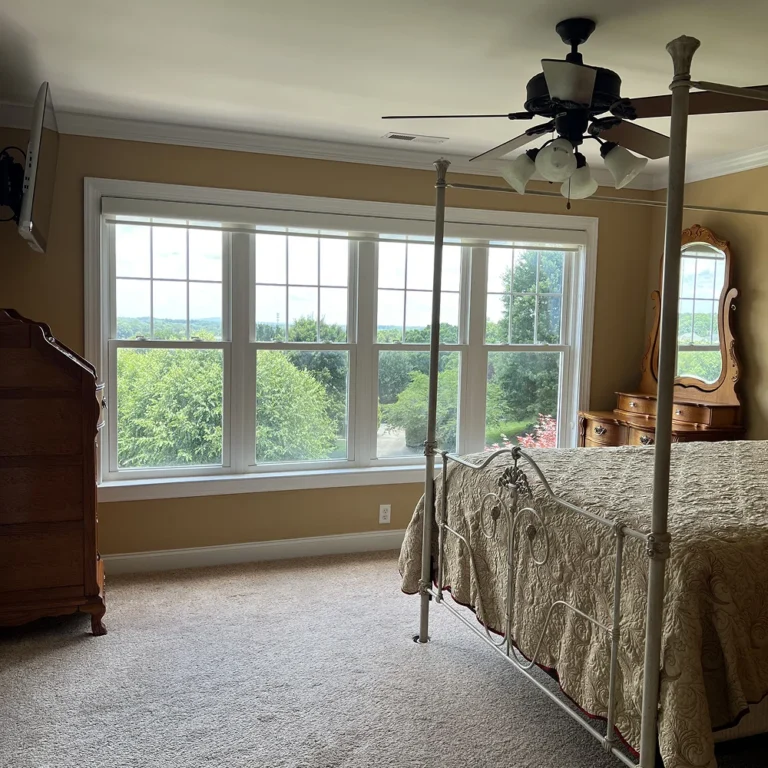A traditional bedroom with beige walls and carpeted flooring, featuring a large white-trimmed window overlooking green trees and hills. The room includes a four-poster bed with a patterned quilt, an antique dresser with a mirror, and a ceiling fan with lights.