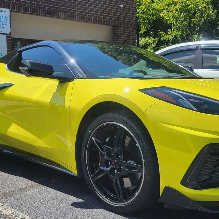 Bright yellow Chevrolet Corvette with dark tinted windows and black rims, parked outside a brick building under direct sunlight.
