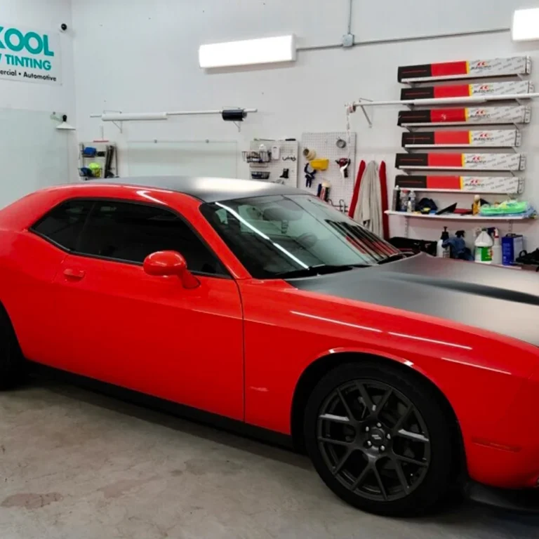 Red Dodge Challenger with dark tinted windows and matte black hood and roof, parked inside Sun Kool Window Tinting shop with tools and film rolls organized on the wall.