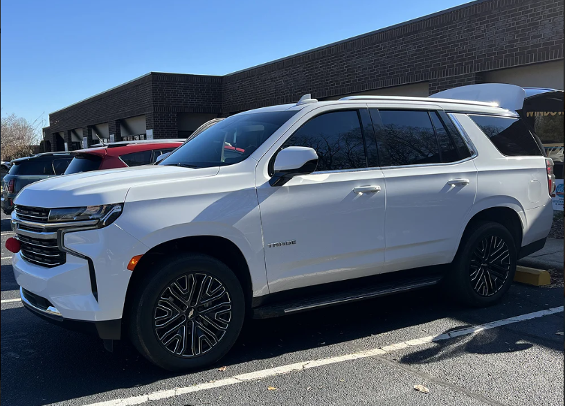 White Chevrolet Tahoe SUV with dark tinted windows and black alloy wheels parked in a shopping center lot on a sunny day.