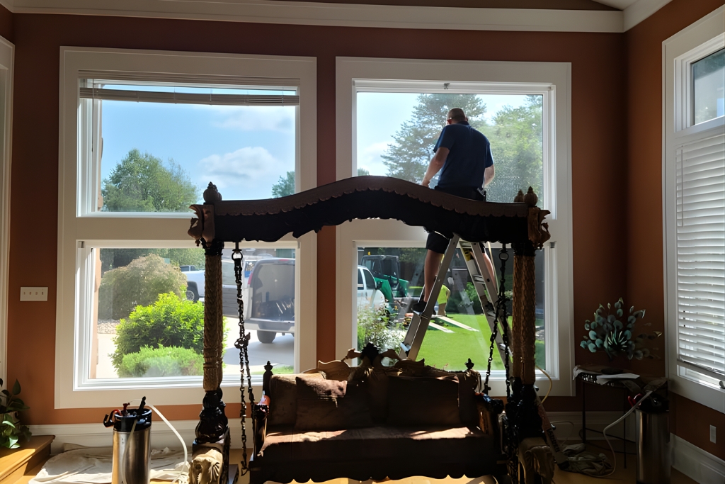 A professional installer applies residential window tinting to a large front-facing window from a ladder, with bright daylight highlighting the contrast between tinted and untinted glass in a well-furnished living room.