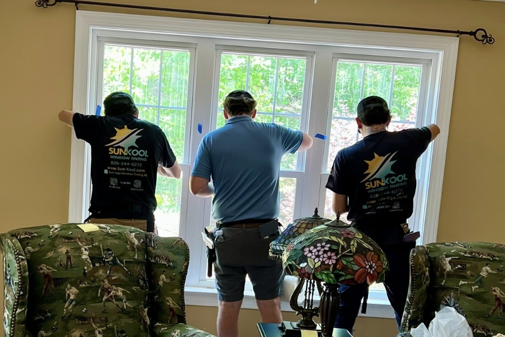 Three professional installers from Sun-Kool Window Tinting apply residential window tinting film to a wide multi-pane window in a living room, demonstrating a precise and team-based approach.