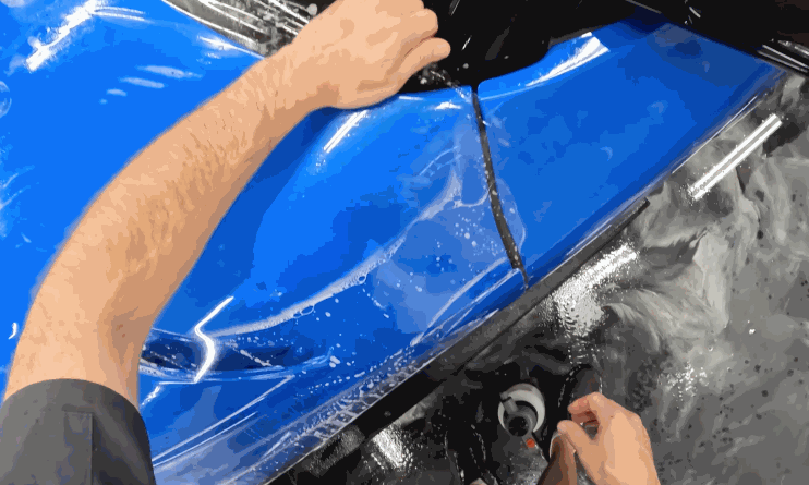Hands stretch and align paint protection film over the front fender of a glossy blue car, with soap solution visible on the surface.