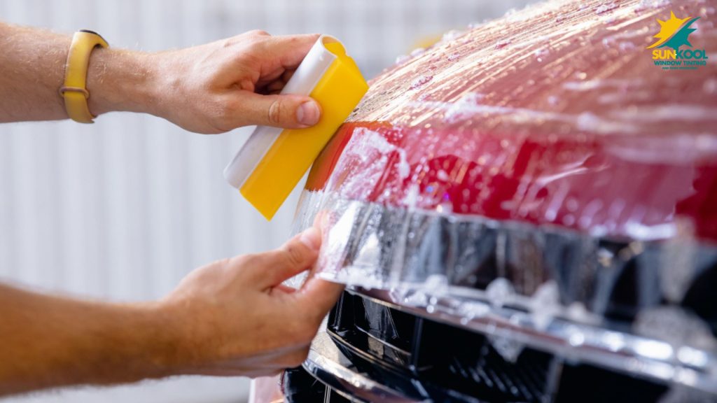 A person uses a yellow squeegee to smooth paint protection film on the curved red front bumper of a car, with the Sun Kool Window Tinting logo visible.