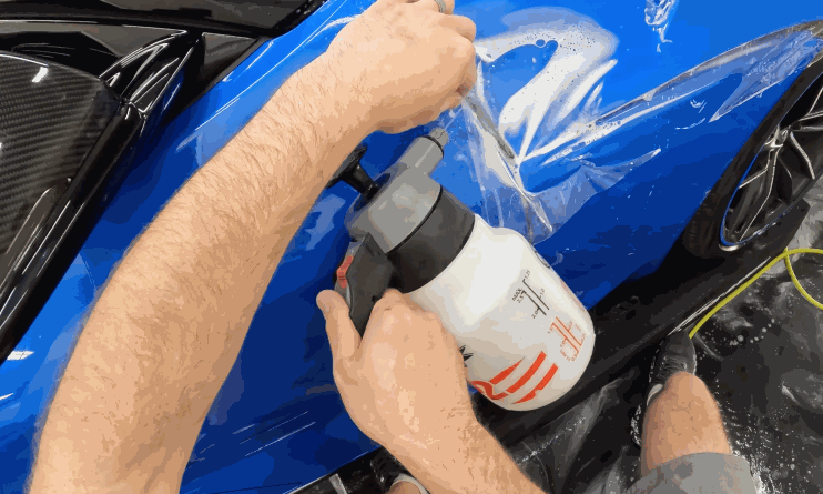 A person applies paint protection film to the side of a blue sports car, using a spray bottle and squeegee to smooth out the clear film.