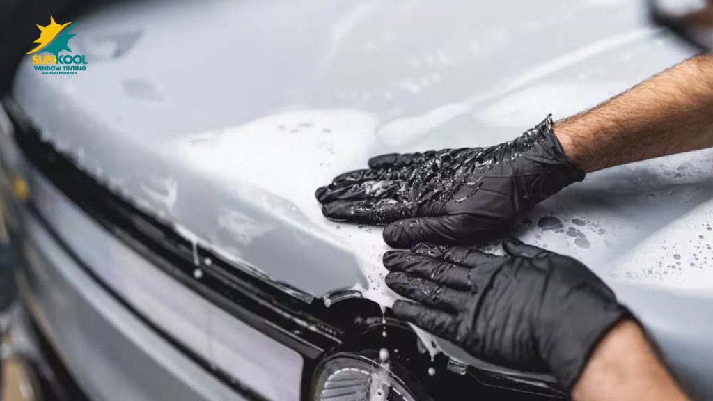 Gloved hands applying paint protection film to a silver car hood using soap and water to ensure a smooth finish.