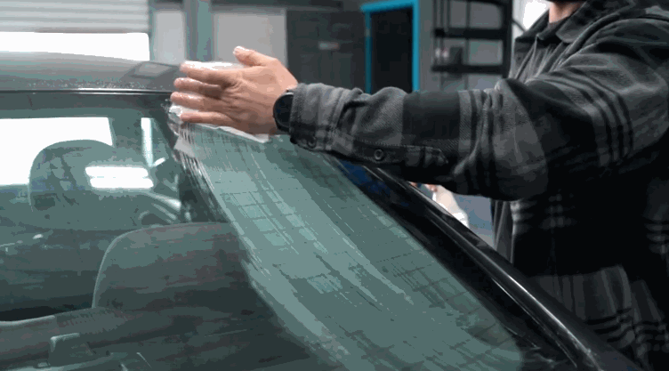 A technician applies a sheet of window tint to the windshield of a car, carefully aligning it across the wet glass surface.