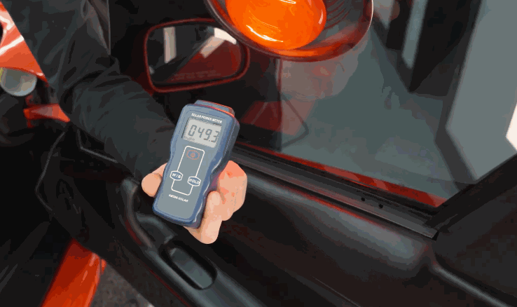 Handheld digital tint meter measures light transmission on a red car window during a window tinting inspection showing a reading of 49.3 percent. The device is held against the driver side window near the side mirror.