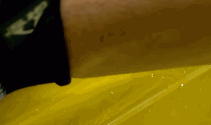 Extreme close up of a glossy yellow car surface with paint protection film being laid down, showing water droplets and slight creases as it is adjusted. The focus highlights the texture and careful placement of the film during installation.