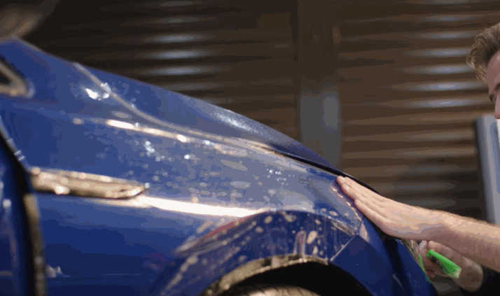 A blue car fender covered in soapy water as a technician’s hand smooths paint protection film over the curved surface. The film reflects light while being carefully aligned and pressed down to remove bubbles.