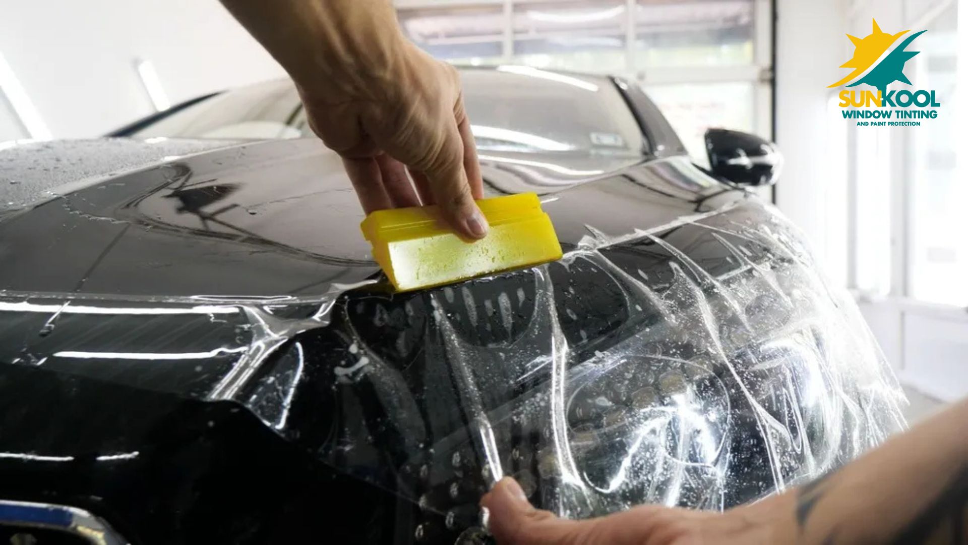 Close up of a black car headlight while a technician uses a yellow squeegee to apply paint protection film smoothly across the surface. Water and bubbles are visible under the film as it is pressed into place in a garage setting. The logo "SUNKOOL WINDOW TINTING AND PAINT PROTECTION" appears in the top right corner.