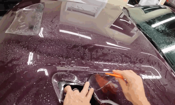 A technician uses an orange squeegee to smooth clear paint protection film over the wet hood of a burgundy car. The film is being shaped around a hood vent for a close custom fit.