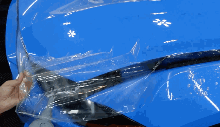 Clear paint protection film is pulled across the front corner of a bright blue car near the headlight. The film is being positioned over the paint to protect the bumper and curved body panels.