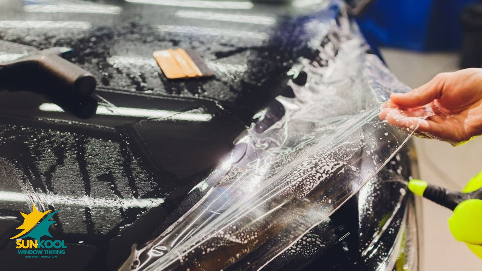 Clear paint protection film being stretched across the wet front end of a black car during installation. A hand smooths the film near the headlight.